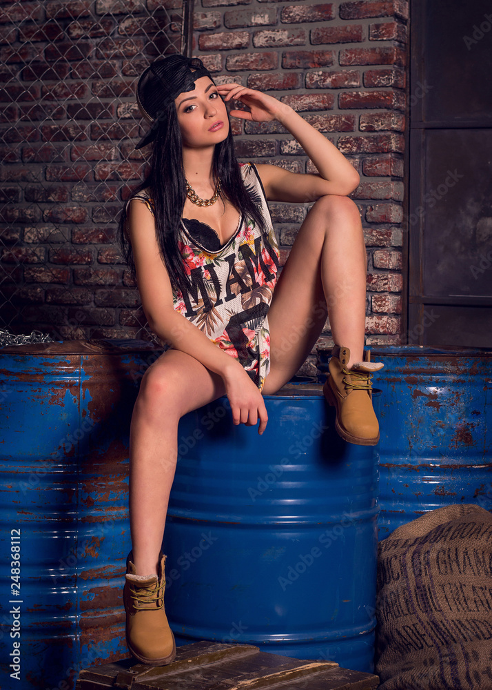 Beautiful tanned sexy girl in short denim shorts. A girl in a T-shirt is  sitting on iron barrels spreading her legs. Conceptual photography for  clothing stores Stock Photo | Adobe Stock