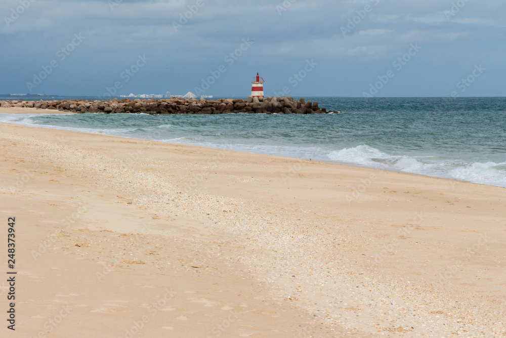 Sandy beach before the rain. Dark clouds over the ocean and the beach. Red and white lighthouse on the rocks.