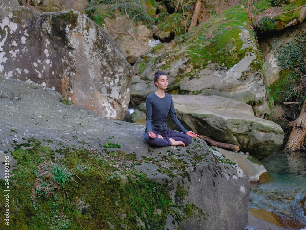 Young slim woman practicing yoga outdoors on big moss rock. Unity with nature concept. Girl meditates in easy pose
