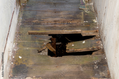 A hole in the floor of an old building. The floor is in a narrow hallway. A dark basement can be seen through the hole.