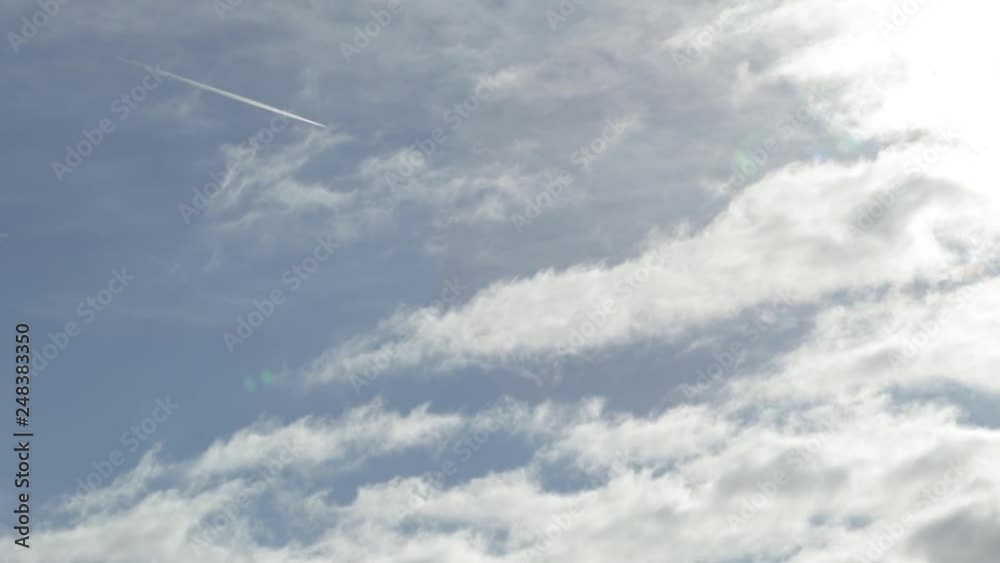 A flying airplane generating a condensation trails (contrail) over the fast-moving clouds and the blue sky in the background. A flying airplane generating a condensation trails (contrail) over the fast-moving clouds and the blue sky in the background.