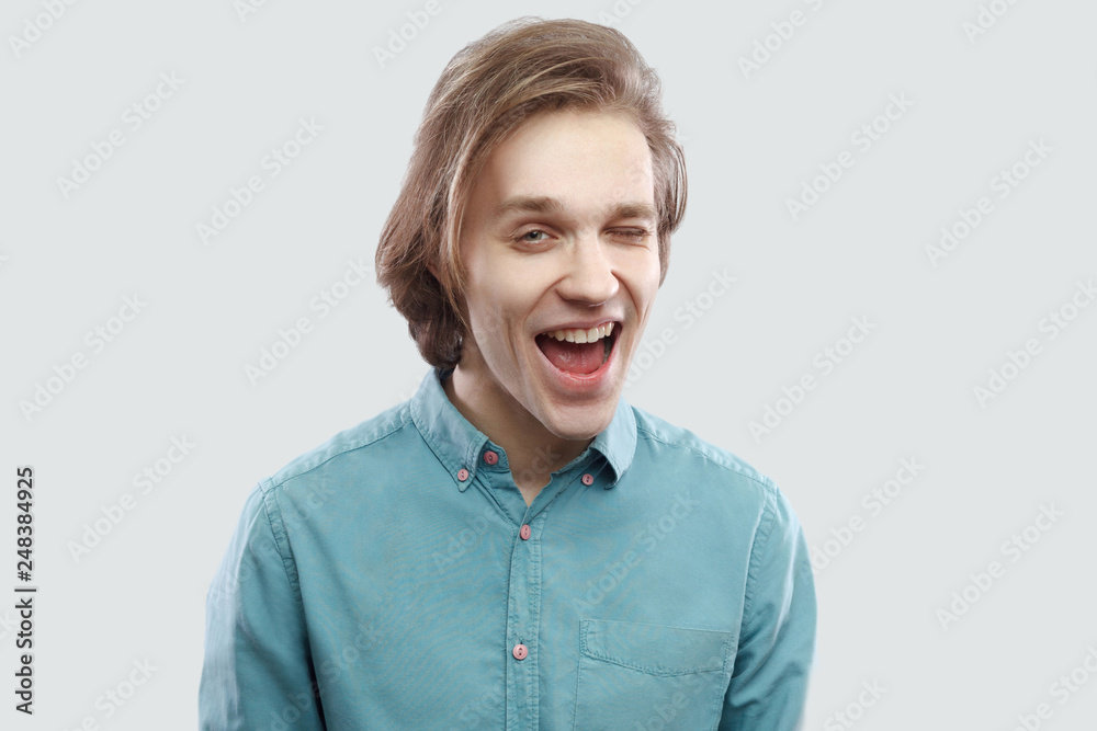 Portrait of funny happy handsome long haired blonde young man in blue casual shirt standing and looking at camera winking. indoor studio shot, isolated on light grey background. Portrait of funny happy handsome long haired blonde young man in blue casual shirt standing and looking at camera winking. indoor studio shot, isolated on light grey background.