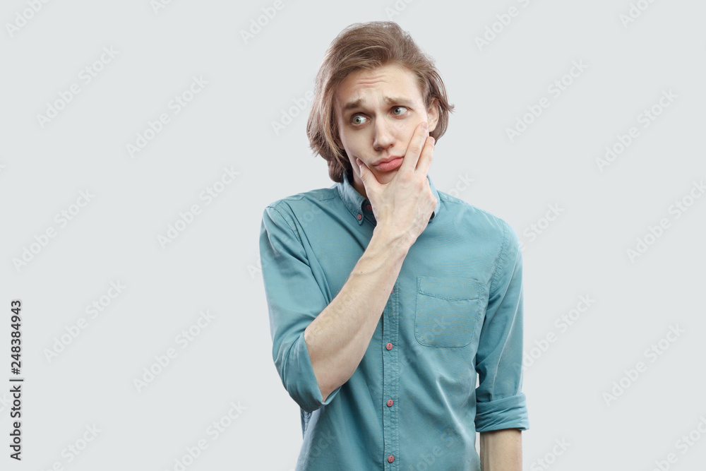 Portrait of thoughtful handsome long haired blonde young man in blue casual shirt standing, thinking, tiuching his face and looking away. indoor studio shot, isolated on light grey background. Portrait of thoughtful handsome long haired blonde young man in blue casual shirt standing, thinking, tiuching his face and looking away. indoor studio shot, isolated on light grey background.