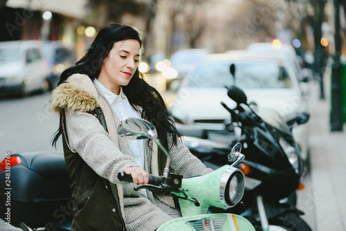 Brunette woman riding on a scooter parked on the street posing.