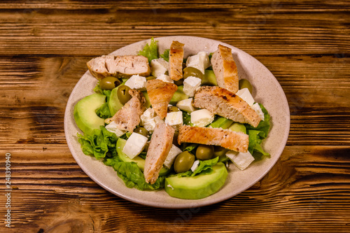 Fresh salad with chicken meat, feta cheese, avocado, green olives and lettuce leaves in ceramic plate on wooden table