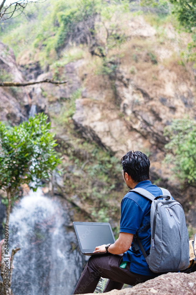 Young man sit and turn back working with laptop at waterfall.