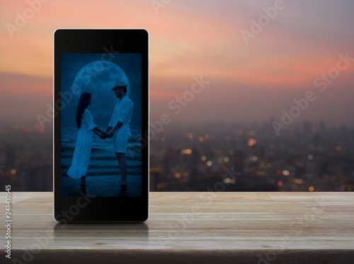 Romantic loving couple standing and holding hands together on modern smart mobile phone screen on wooden table over blur of cityscape on warm light sundown, Business internet dating online, Valentines