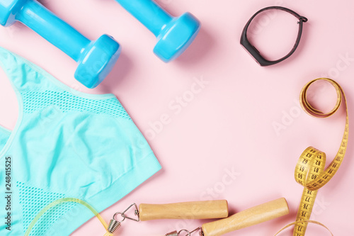 Sport bra, dumbbells, measuring tape, jump rope and fitness tracker on a pink background, top view with copy space. Fitness and body care concept