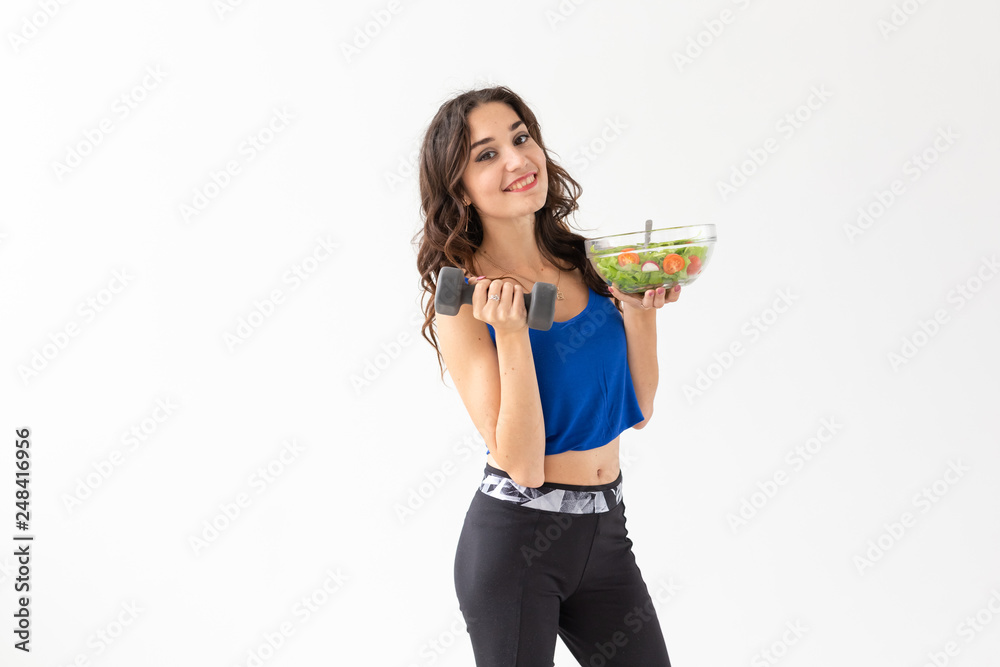 Healthy lifestyle, diet, fitness and people concept - young woman is eating a salad and doing sport with dumbbell on white background Healthy lifestyle, diet, fitness and people concept - young woman is eating a salad and doing sport with dumbbell on white background