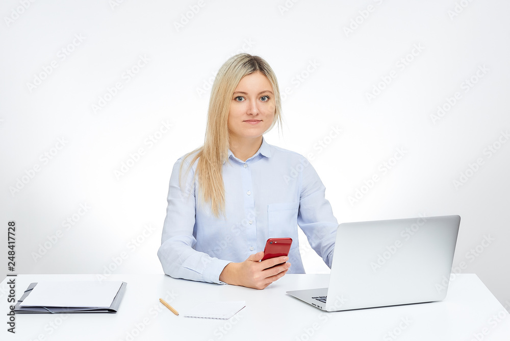 Young blonde woman watches straight holds her red glass cell phone works with her aluminum notebook computer and sits by the desk in the office with the white background. Documents on the table. Young blonde woman watches straight holds her red glass cell phone works with her aluminum notebook computer and sits by the desk in the office with the white background. Documents on the table.