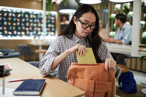 Teenage girl putting her notebook and books into backpack while going to leave college cafe