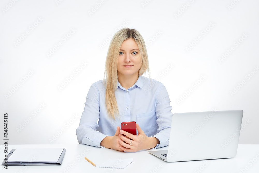 Young blonde woman watches straight holds her red glass cell phone and sits by the desk in the office with the white background. Aluminum notebook computer and documents on the table. Young blonde woman watches straight holds her red glass cell phone and sits by the desk in the office with the white background. Aluminum notebook computer and documents on the table.