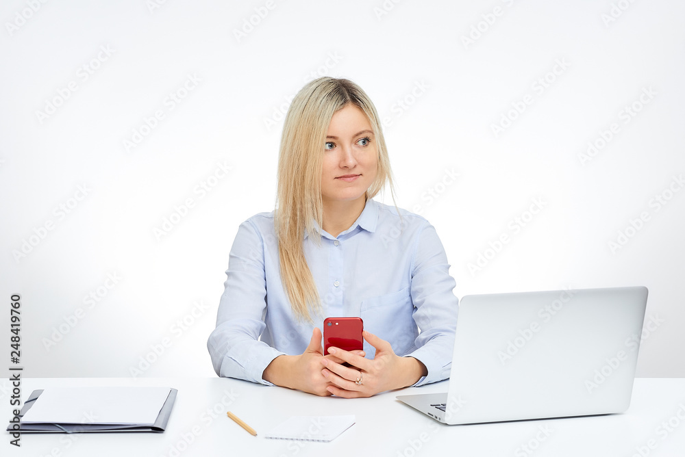 Young blonde woman looks away holds her red glass cell phone and sits by the desk in the office with the white background. Aluminum notebook computer and documents on the table. Young blonde woman looks away holds her red glass cell phone and sits by the desk in the office with the white background. Aluminum notebook computer and documents on the table.