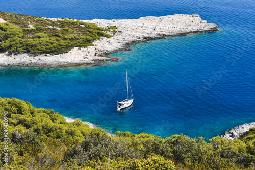 View of a beautiful blue bay with a white sailing boat anchored in clear blue and turquoise transparent water with view on white rocks and green mediterranean vegetation. Summer scene from Croatia