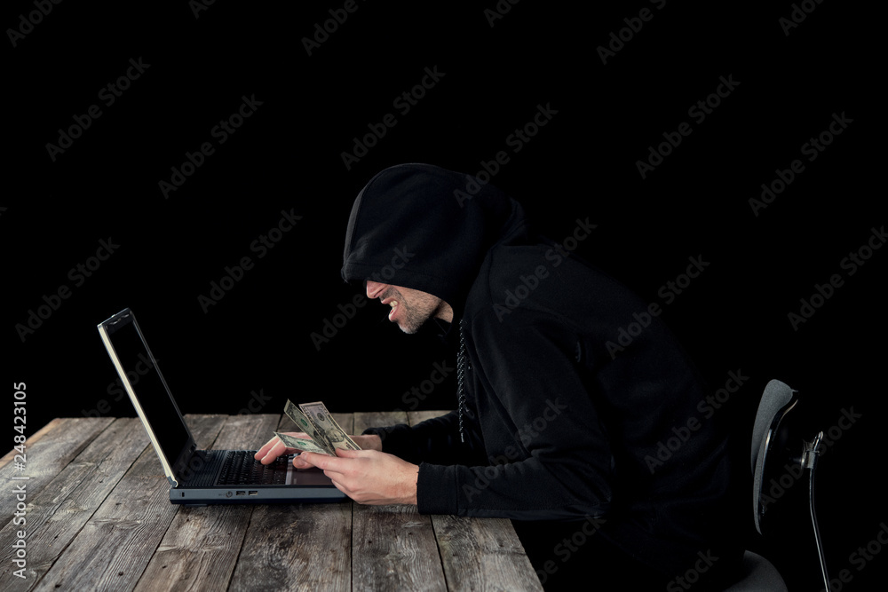 hacker in black hoody with laptop and dollar notes hacker in black hoody with laptop and dollar notes