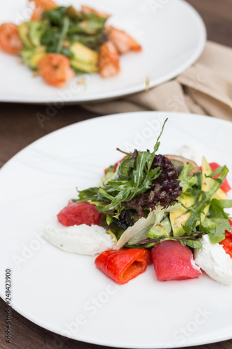 A salad with red fish and greens on a white plate is on the table against the background of another salad.