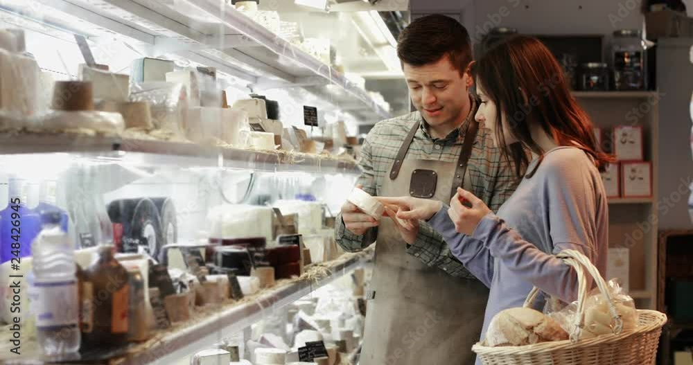 Male Sales Assistant Giving Advice To Female Customer In Delicatessen Shopping For Cheese