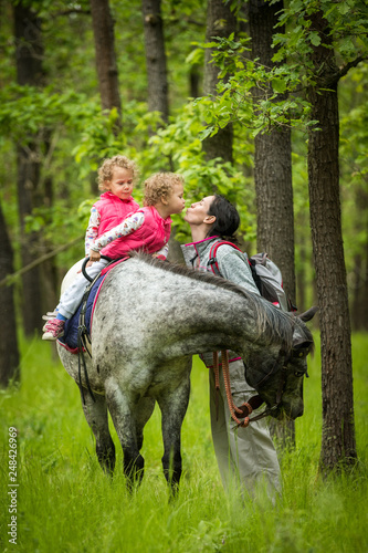 Girls enjoying horseback riding in the woods and kissing mother, young pretty girls with blond curly hair on a horse, freedom, joyful, outdoor, spring, healthy, happy hamily, kids on vacation forest © Ji