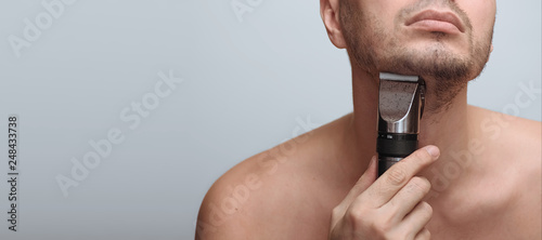 Cropped image of a young bearded man using electric shaver isolated over gray background