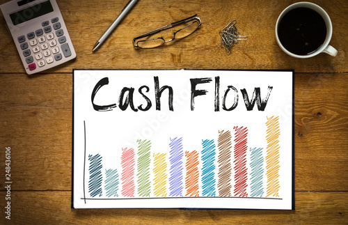 Overhead top view of Cash Flow written in book on desk with a coloured bar chart and trend line.  Desk has coffee cup, glasses, pen and calculator photo