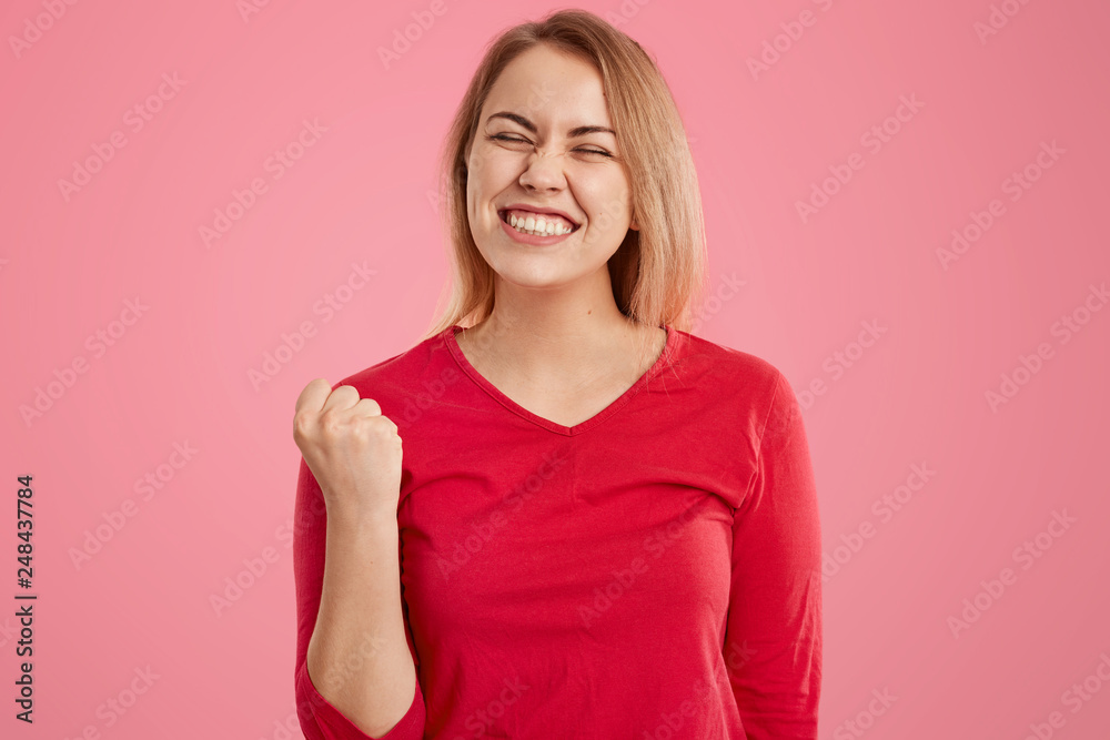 Overjoyed European woman with short hair, clenches fist, dressed in red casual outfit, squints face, dressed in red jumper, isolated over pink background. Female winner rejoices success or triumph Overjoyed European woman with short hair, clenches fist, dressed in red casual outfit, squints face, dressed in red jumper, isolated over pink background. Female winner rejoices success or triumph