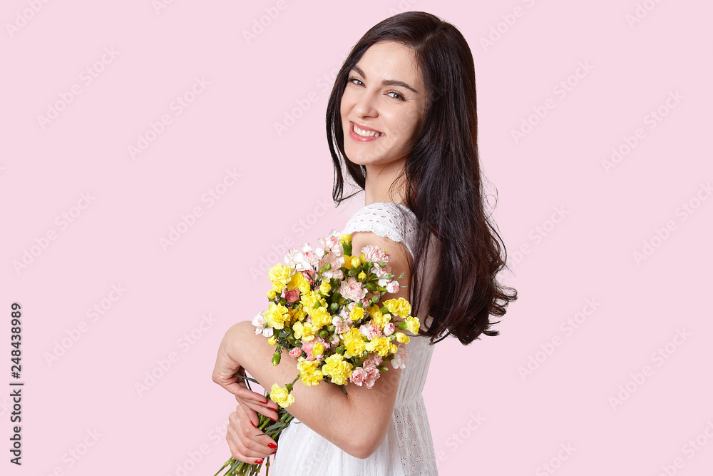 Photo of attractive brunette young woman with pleasant smile, dressed in white dress, holds bouquet of flowers, looks directly at camera, poses over light pink background, has minimal make up Photo of attractive brunette young woman with pleasant smile, dressed in white dress, holds bouquet of flowers, looks directly at camera, poses over light pink background, has minimal make up
