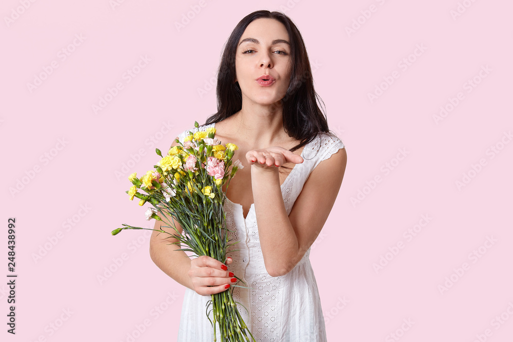 Brunette lovely woman with dark hair, appealing look, blows air kiss, flirts with boyfriend on distance, holds flowers, dressed in summer white dress, isolated over rosy studio wall. You are my love Brunette lovely woman with dark hair, appealing look, blows air kiss, flirts with boyfriend on distance, holds flowers, dressed in summer white dress, isolated over rosy studio wall. You are my love