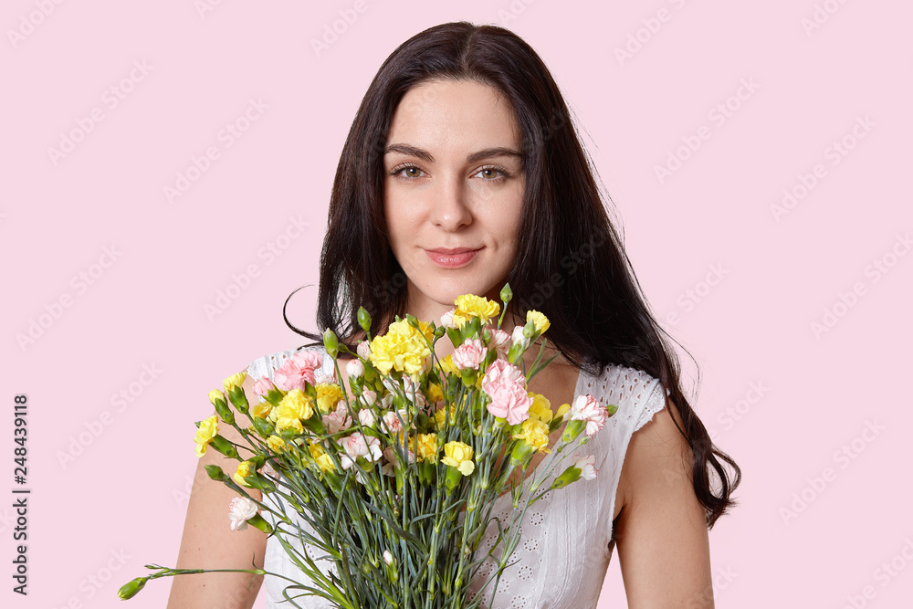 Close up shot of lovely brunette young woman with healthy soft skin, minimal make up, holds flowers in front, looks at camera with happiness, poses over rosy background indoor. Women and flowers