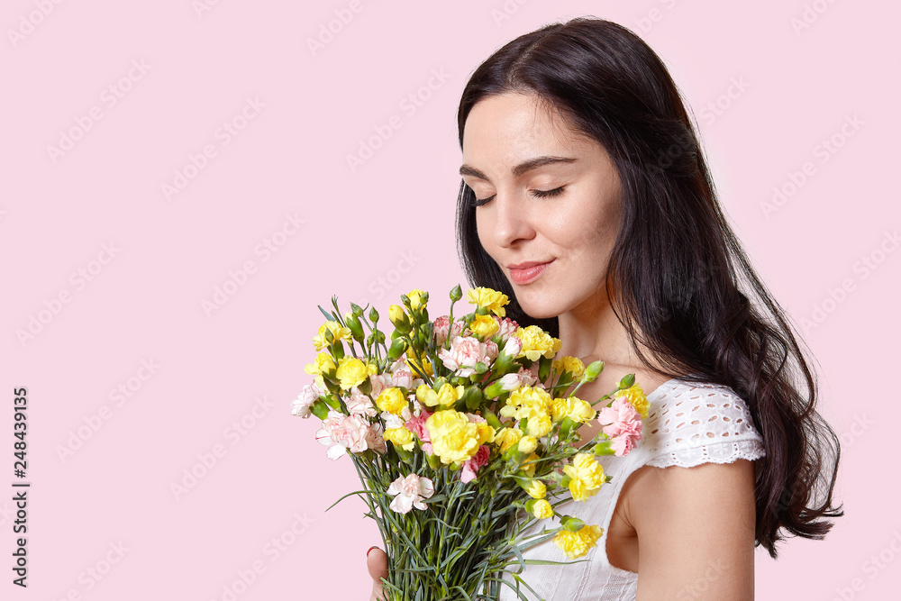 Image of attractive woman with pleasant look, closes eyes, smells flowers, celebrates International Womens Day, dressed in white dress, isolated over pink wall with empty space for information Image of attractive woman with pleasant look, closes eyes, smells flowers, celebrates International Womens Day, dressed in white dress, isolated over pink wall with empty space for information