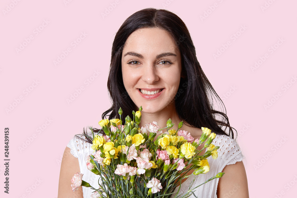 Photo of pleasant looking dark haired European woman smiles sensually, has soft skin, enjoys good day, recieves bouquet of spring flowers, has natural beauty, isolated over rosy studio wall.