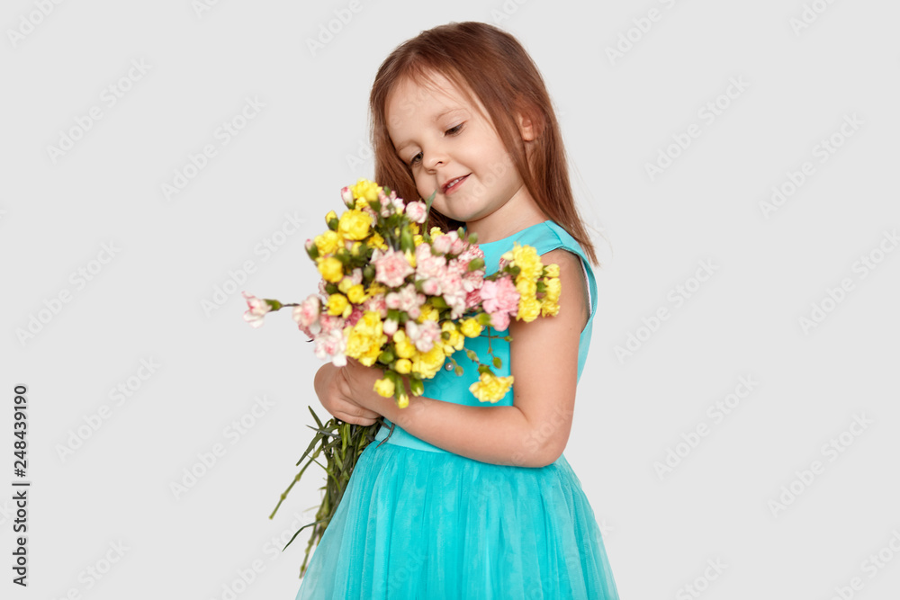 Sideways shot of attractive small child with long straight hair, dressed in blue stylish dress, looks positively at beautiful bouquet of flowers, isolated over white background. Children concept Sideways shot of attractive small child with long straight hair, dressed in blue stylish dress, looks positively at beautiful bouquet of flowers, isolated over white background. Children concept
