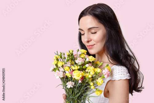 Image of attractive woman with pleasant look, closes eyes, smells flowers, celebrates International Womens Day, dressed in white dress, isolated over pink wall with empty space for information