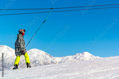 Snowboarder in equipment rises on a lift uphill for skiing against a blue sky and mountain peaks.