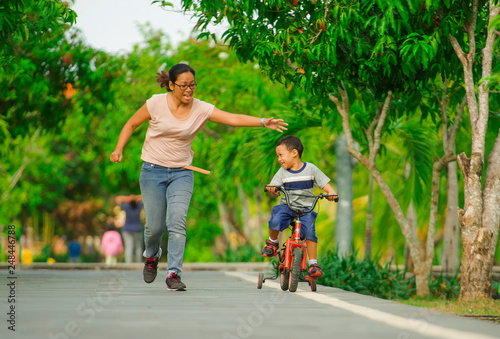 lifestyle portrait of Asian Indonesian mother and young happy son at city park having fun together the kid learning bike riding and the woman running after the child