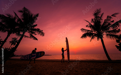 Photography Silhouette of surfers couple holding long surf boards at sunset on tropical beac