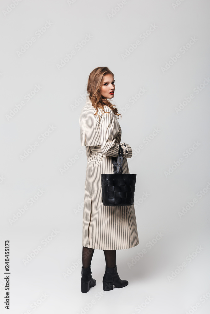 Young attractive woman with wavy hair in striped coat holding black bag dreamily looking in camera standing from back over gray background Young attractive woman with wavy hair in striped coat holding black bag dreamily looking in camera standing from back over gray background
