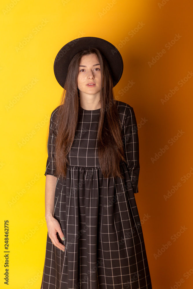 brunette girl is dressed in a stylish dark dress and a wide dark Fedora hat slides for a photo in a photo studio on a yellow background near the windows. Talented charismatic model brunette girl is dressed in a stylish dark dress and a wide dark Fedora hat slides for a photo in a photo studio on a yellow background near the windows. Talented charismatic model