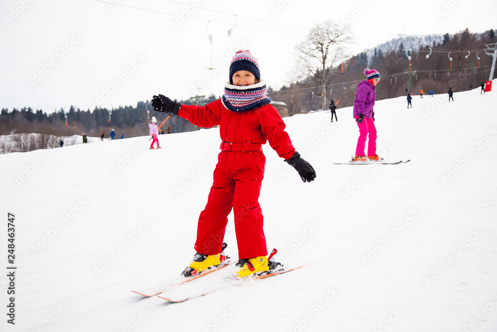 Little smiling girl in red overall training skiing