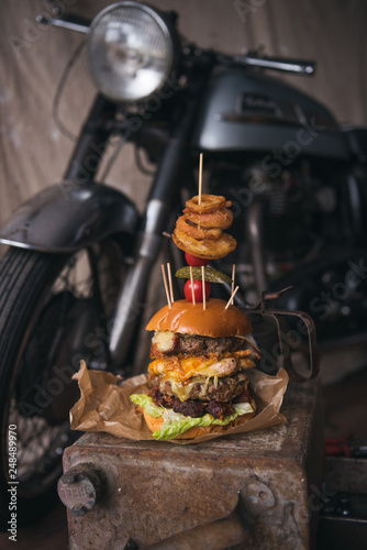 A huge stacked double cheeseburger, with bacon, cheese, lettuce, topped with gherkins, onion rings and cherry tomato. photographed in a rustic mechanic workshop background. © dannyburn