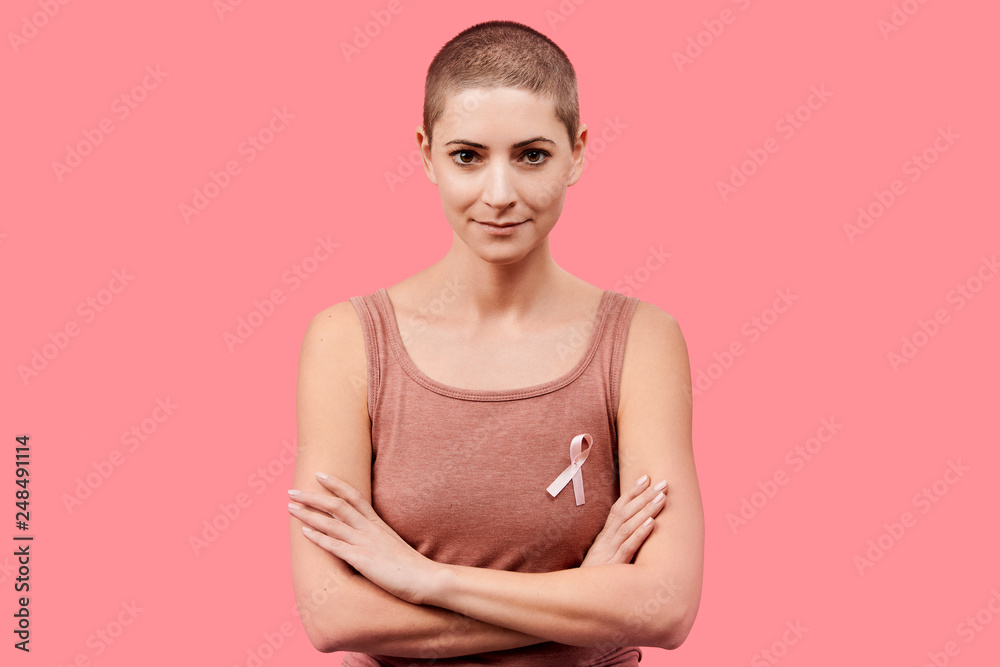Obraz premium Smiling mid 30s woman, a cancer survivor, wearing pink breast cancer awareness ribbon, isolated over living coral background. Support, solidarity, screening and prevention concept.