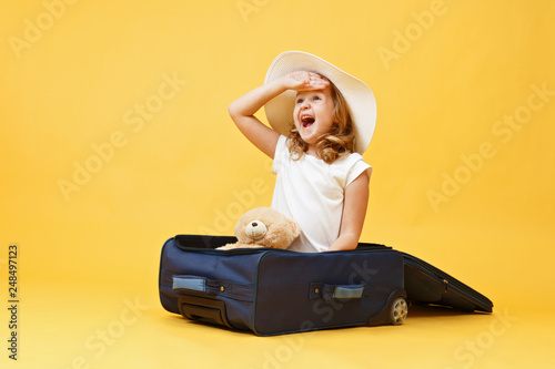 A little blonde child girl dreams of traveling and sits in a suitcase with a toy bear.Yellow background