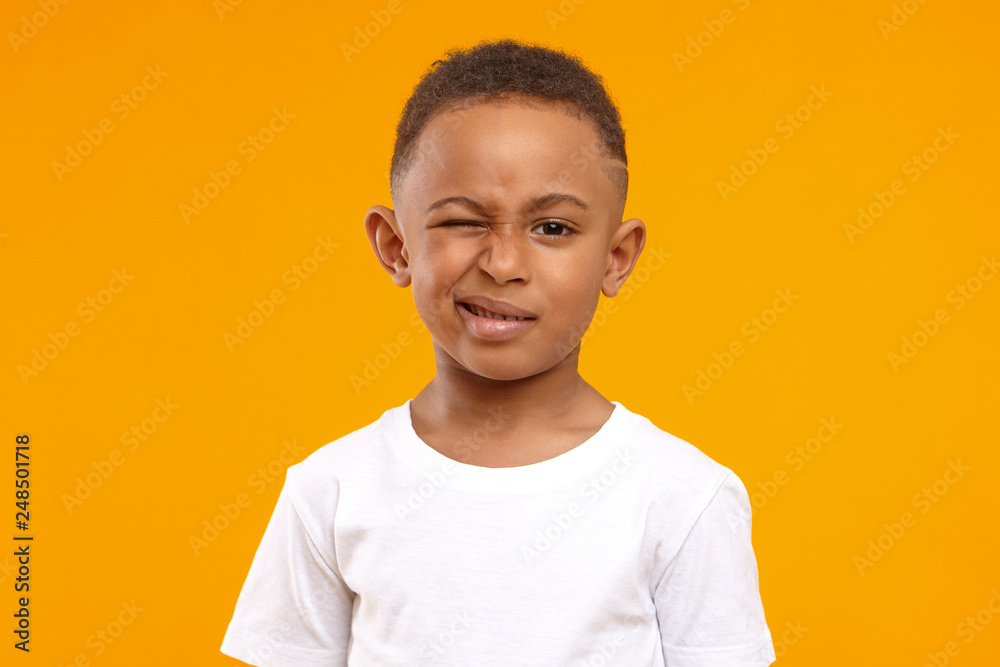 Children, fun and body language concept. Cute African American little boy grimacing against yellow studio wall background, winking at camera, planning joke or trick, wearing casual white t-shirt