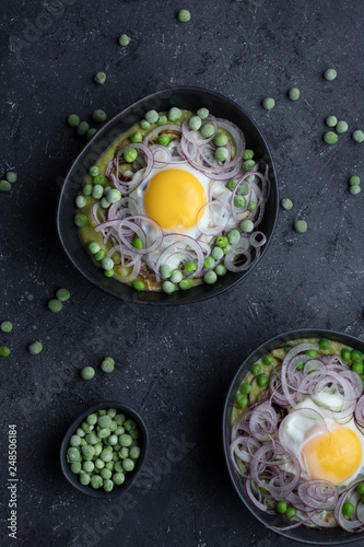 Two bowls with green pea soup, fried egg and red onion on dark background, top view.