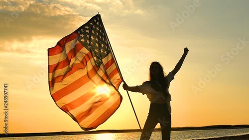 Long hair girl  patriotic teenager  stand with American flag against sky with  orange sun. Slow motion