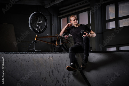Young handsome BMX rider relaxing after practicing tricks, sitting on a floor in a skatepark indoors