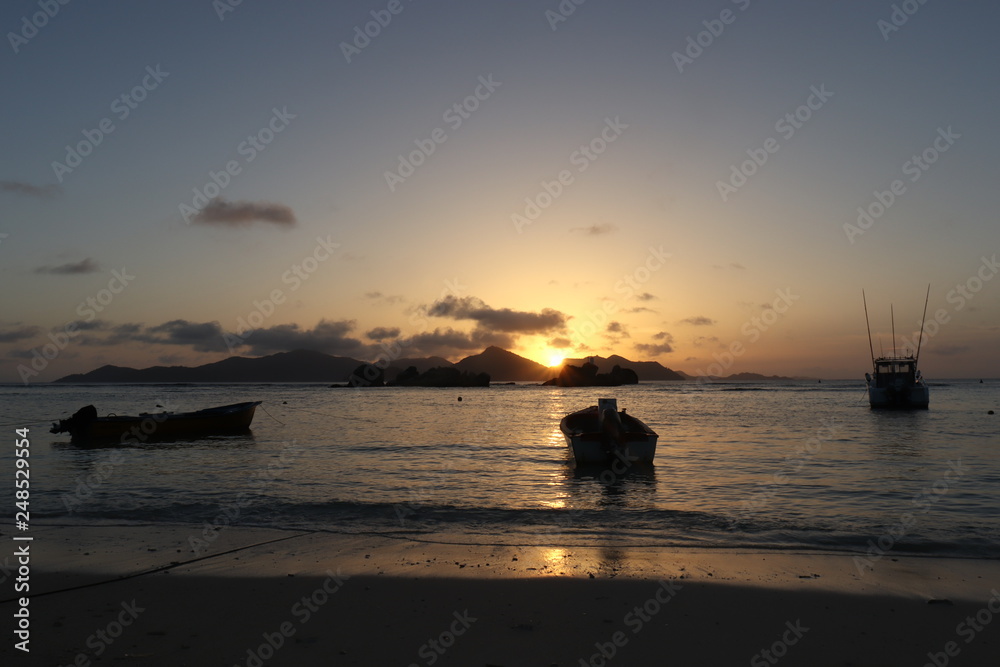Tropical sunset at beach. Anchored boats in the ocean and the Island of Praslin.