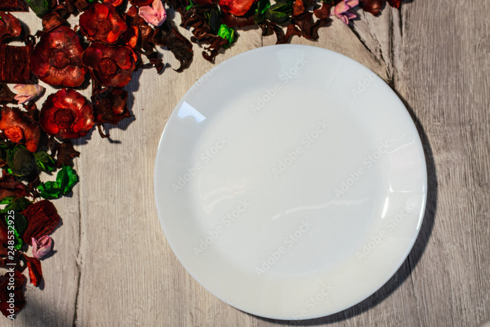 Empty white plate on a wooden background decorated with dry flower petals. View from above. Empty white plate on a wooden background decorated with dry flower petals. View from above.