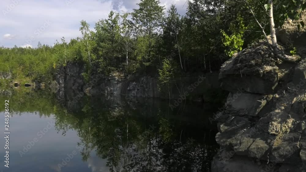 Stony shore at the lake. A young mixed forest grows on a high rocky shore of a lake. Small area with green grass and moss. Small lake in nature Stony shore at the lake. A young mixed forest grows on a high rocky shore of a lake. Small area with green grass and moss. Small lake in nature