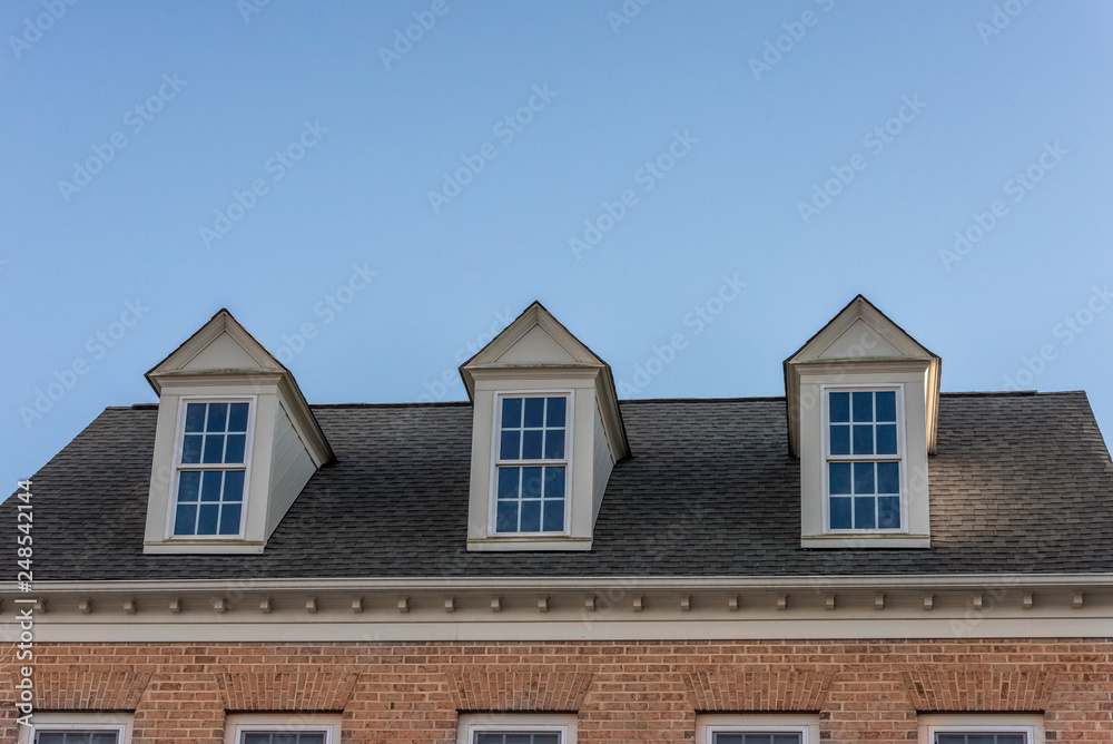Three white dormer sash window on brick facade wall with blue sky background on a gable roof with vinyl siding on a luxury estate house in the East Coast of the USA for upper middle class families Three white dormer sash window on brick facade wall with blue sky background on a gable roof with vinyl siding on a luxury estate house in the East Coast of the USA for upper middle class families