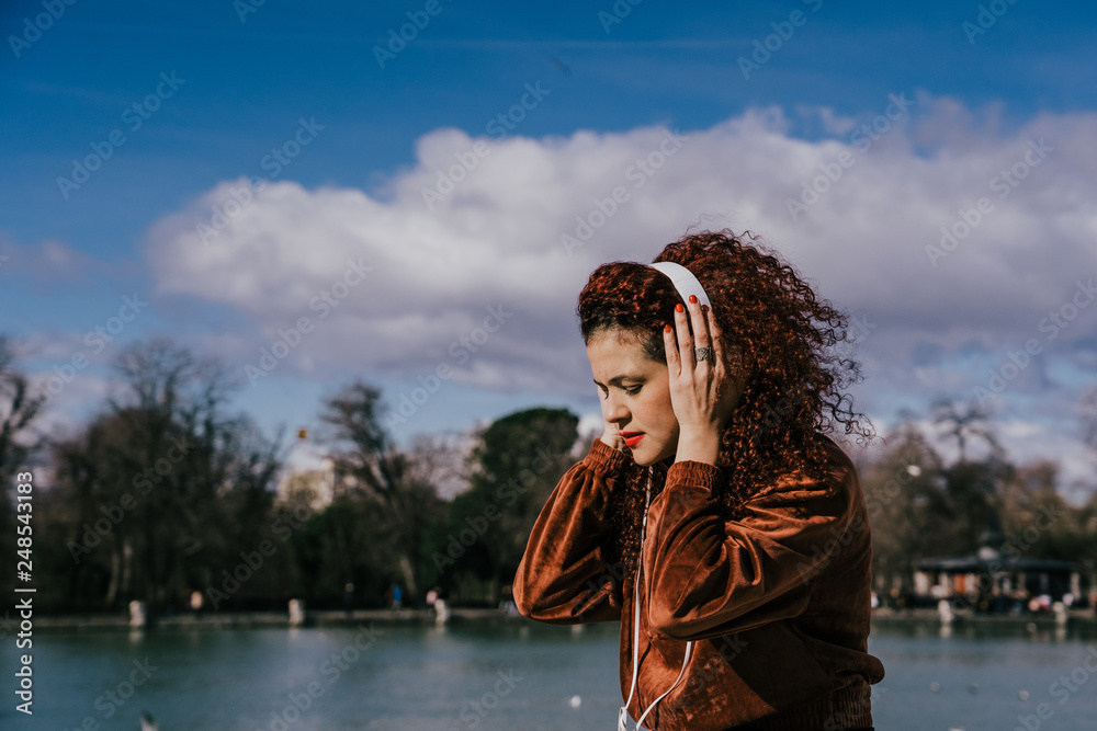 Young woman with afro hair listening to music with headphones at the edge of a small lake. Full of positive energy, fun and carefree. Lifesytle.